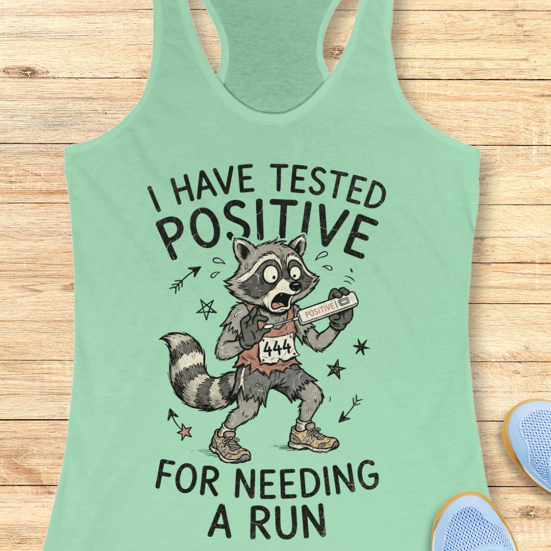 A mint green racerback tank top laid flat on a light wood background, featuring a cartoon raccoon wearing a race bib and holding a pill bottle, with the text 'I HAVE TESTED POSITIVE FOR NEEDING A RUN'. Partial blue shoes are visible at the bottom corners.