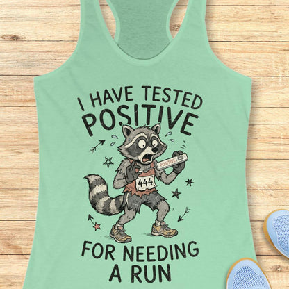 A mint green racerback tank top laid flat on a light wood background, featuring a cartoon raccoon wearing a race bib and holding a pill bottle, with the text 'I HAVE TESTED POSITIVE FOR NEEDING A RUN'. Partial blue shoes are visible at the bottom corners.