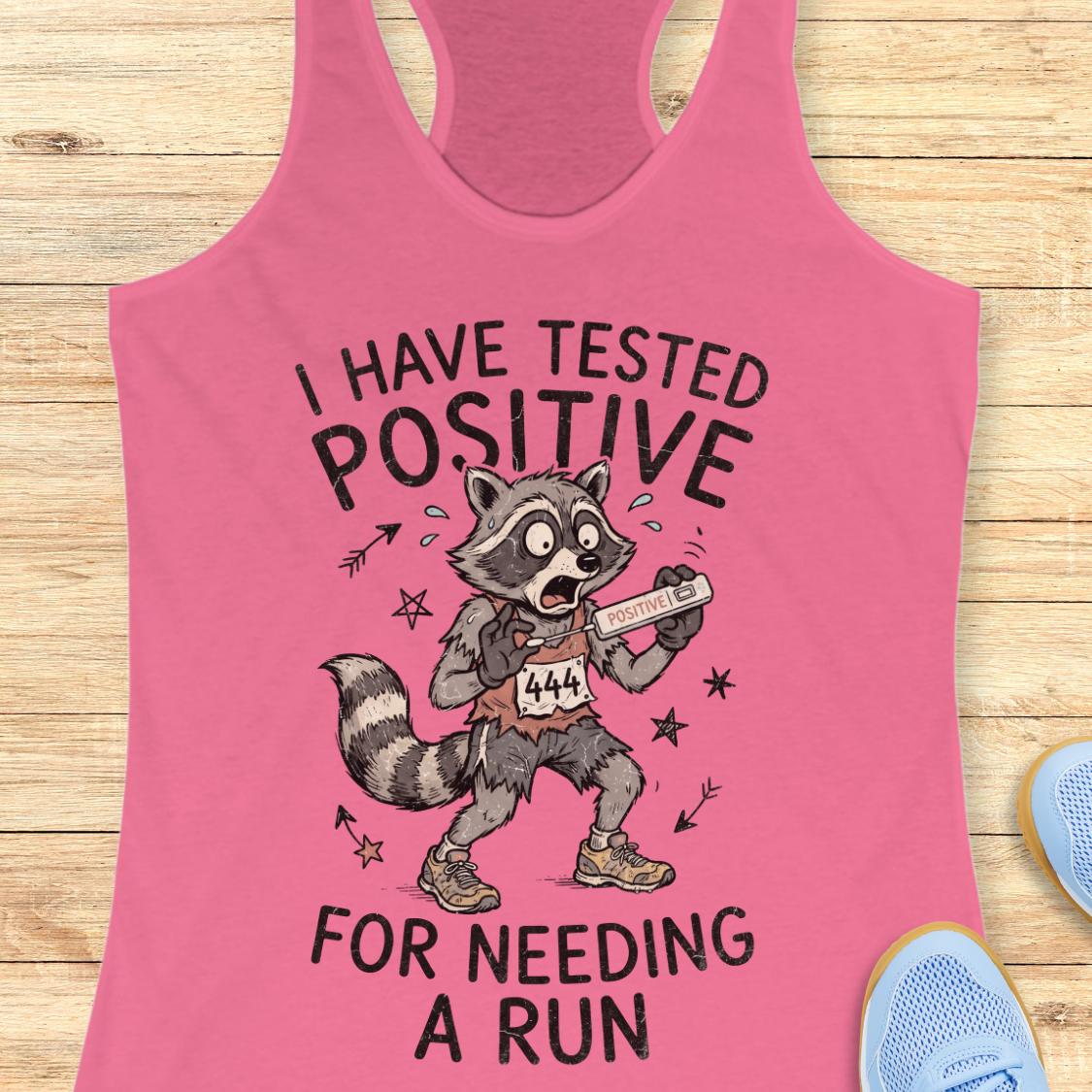 A pink racerback tank top laid flat on a light wood surface, featuring a cartoon raccoon holding a race bib with the text 'I HAVE TESTED POSITIVE FOR NEEDING A RUN.' Partial blue shoes are visible at the edges as styling props.