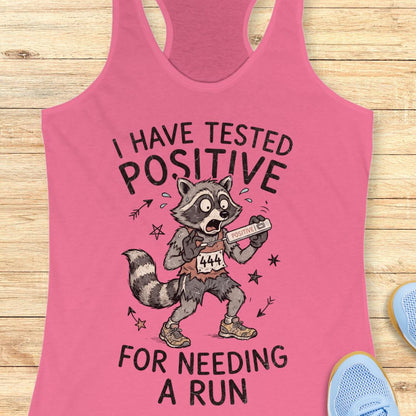 A pink racerback tank top laid flat on a light wood surface, featuring a cartoon raccoon holding a race bib with the text 'I HAVE TESTED POSITIVE FOR NEEDING A RUN.' Partial blue shoes are visible at the edges as styling props.