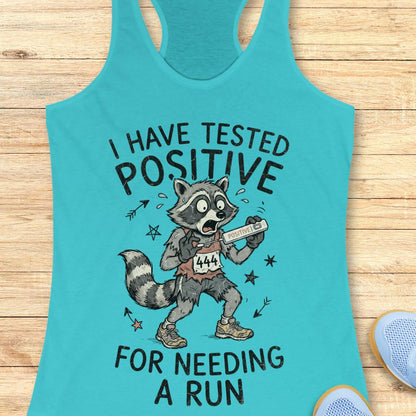 A turquoise racerback tank top laid flat on a light wood surface, featuring a humorous graphic of a raccoon runner holding a bib number 444 with the text 'I HAVE TESTED POSITIVE FOR NEEDING A RUN'.