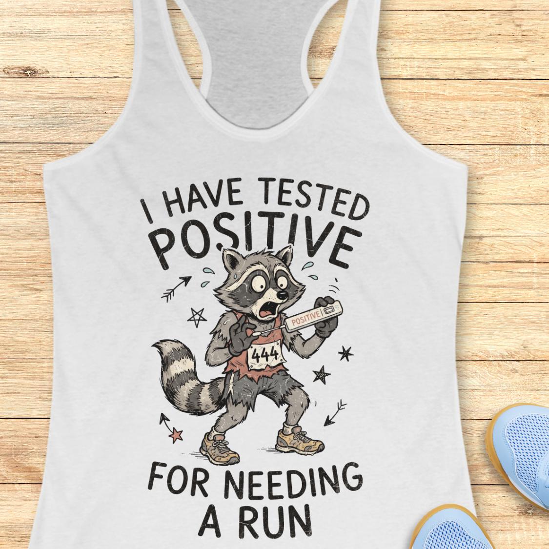 A white racerback tank top laid flat on a light wood background, featuring a humorous graphic of a raccoon runner with the text 'I HAVE TESTED POSITIVE FOR NEEDING A RUN'.