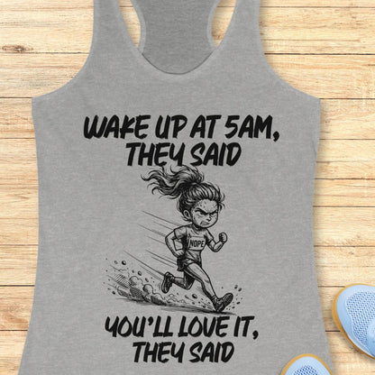 A heather gray racerback tank top laid flat on a light wood surface, featuring a black graphic of a running cartoon woman and the text 'WAKE UP AT 5AM, THEY SAID YOU'LL LOVE IT, THEY SAID'. Partial blue shoes are visible at the bottom right as styling props.