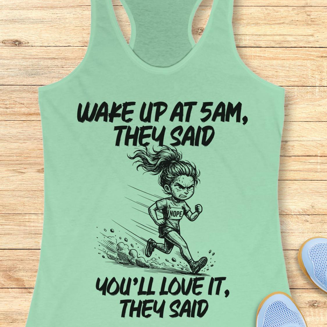 A mint green racerback tank top laid flat on a light wood background, featuring a black cartoon illustration of a runner and the text 'WAKE UP AT 5AM, THEY SAID YOU'LL LOVE IT, THEY SAID'. Partial blue sneakers are visible at the bottom right as styling props.