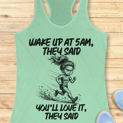 A mint green racerback tank top laid flat on a light wood background, featuring a black cartoon illustration of a runner and the text 'WAKE UP AT 5AM, THEY SAID YOU'LL LOVE IT, THEY SAID'. Partial blue sneakers are visible at the bottom right as styling props.
