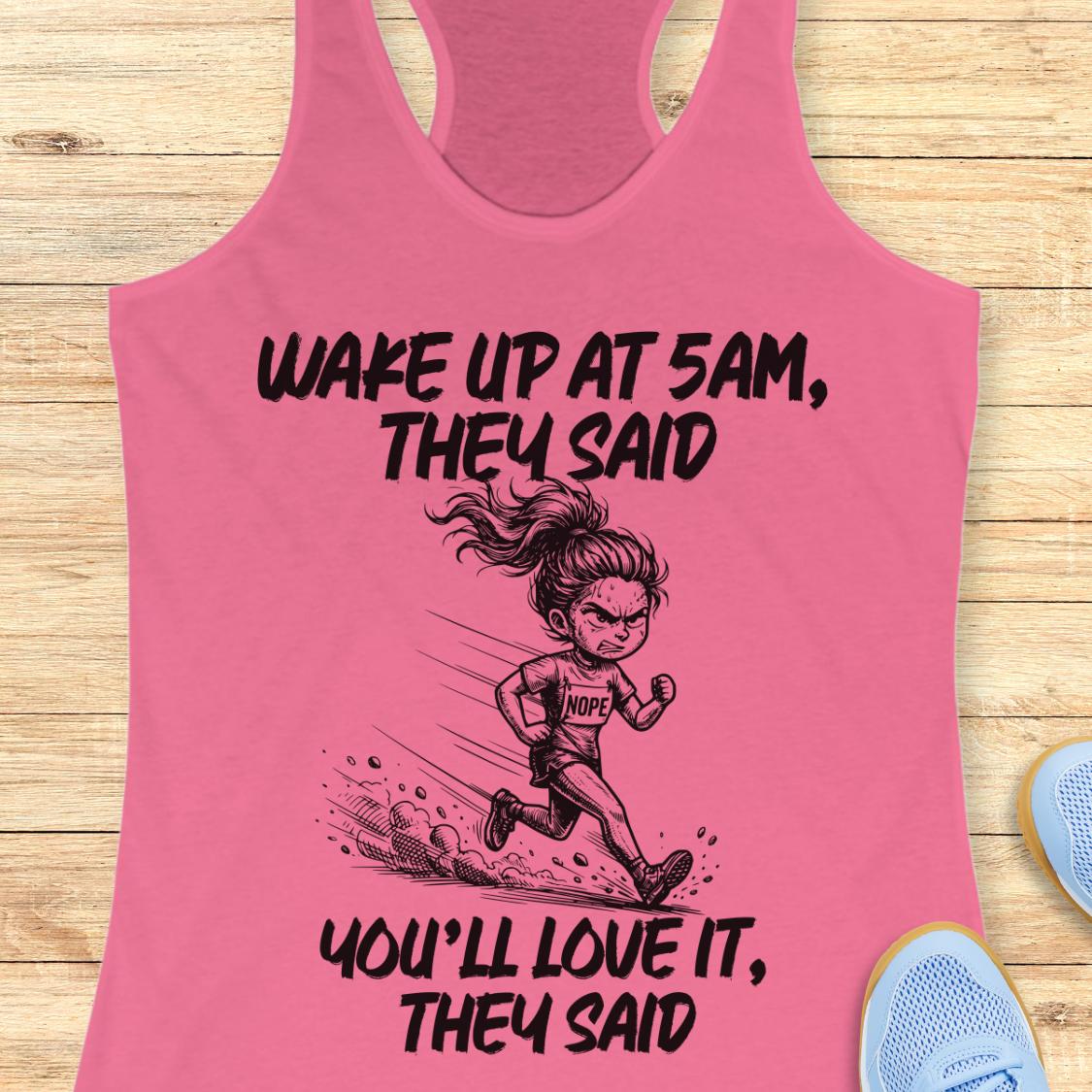 A pink racerback tank top laid flat on a light wood surface, featuring a black graphic of a running woman and the text 'WAKE UP AT 5AM, THEY SAID YOU'LL LOVE IT, THEY SAID'. Partial blue sneakers are visible at the bottom right as styling props.