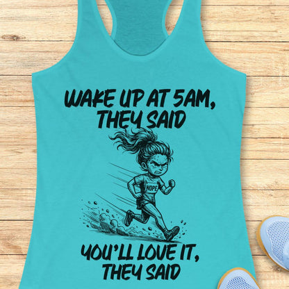 A turquoise racerback tank top laid flat on a light wood surface, featuring a black graphic of a running woman and the text 'WAKE UP AT 5AM, THEY SAID YOU'LL LOVE IT, THEY SAID'. A partial white sneaker is visible at the bottom right.
