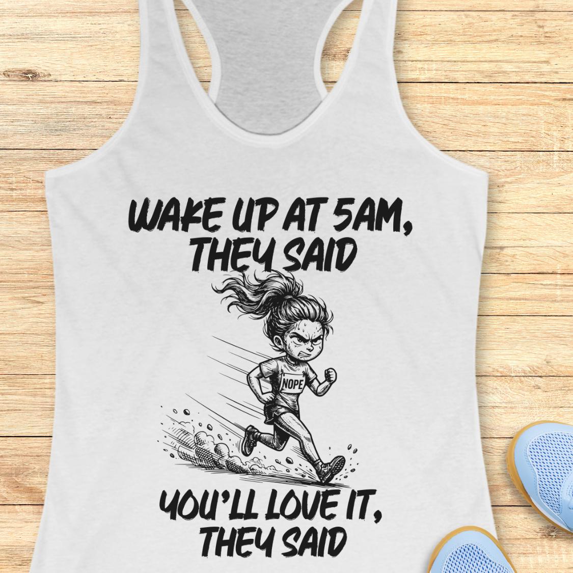 A white racerback tank top laid flat on a light wood background, featuring a black graphic of a running woman and the text 'WAKE UP AT 5AM, THEY SAID YOU'LL LOVE IT, THEY SAID'. Partial blue shoes are visible at the bottom right as styling props.