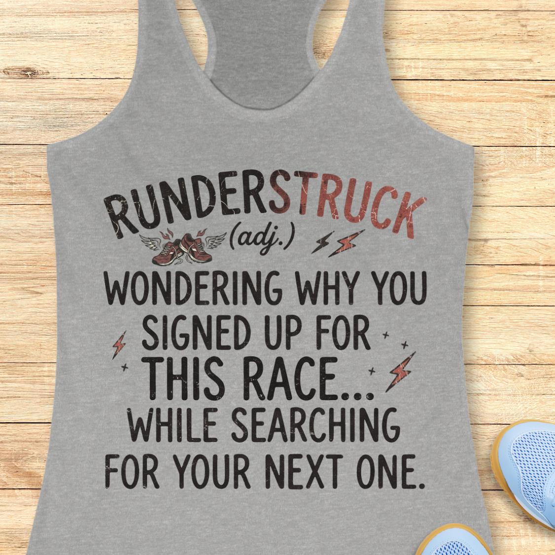 A heather gray racerback tank top laid flat on a light wood surface, featuring a humorous race-themed graphic that reads 'RUNDERSTRUCK (adj.) WONDERING WHY YOU SIGNED UP FOR THIS RACE.