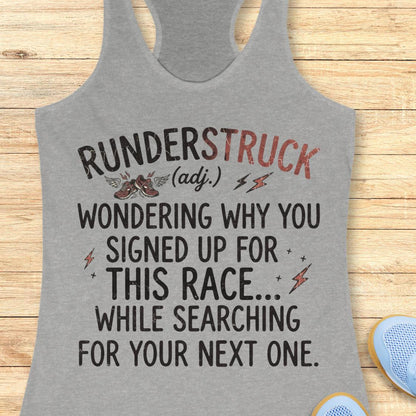 A heather gray racerback tank top laid flat on a light wood surface, featuring a humorous race-themed graphic that reads 'RUNDERSTRUCK (adj.) WONDERING WHY YOU SIGNED UP FOR THIS RACE.