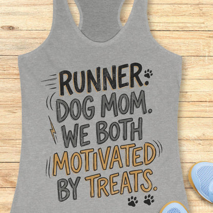 A heather gray racerback tank top laid flat on a light wood background, featuring the printed phrase 'RUNNER DOG MOM WE BOTH MOTIVATED BY TREATS' in black and mustard yellow lettering with small paw print and lightning bolt accents. Partial blue shoes are visible at the bottom right as styling props.