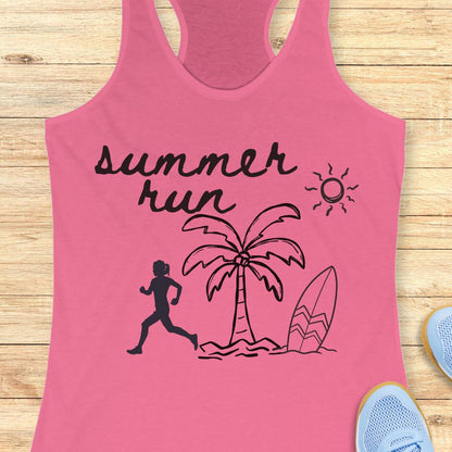 A pink racerback tank top laid flat on a light wood surface, featuring a black graphic that reads 'summer run' with a running figure, palm tree, surfboard, and sun. Partial blue sneakers are visible at the bottom right as styling props.