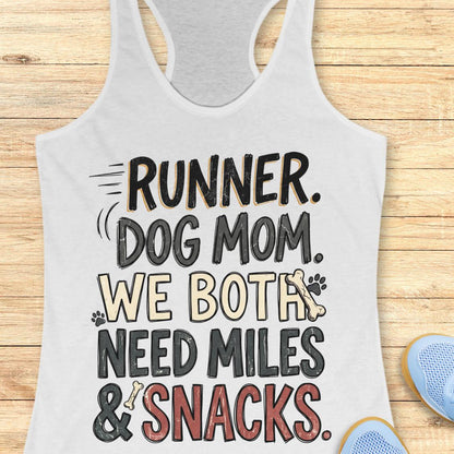 A white racerback tank top laid flat on a light wood background, featuring a humorous graphic that reads 'RUNNER. DOG MOM.