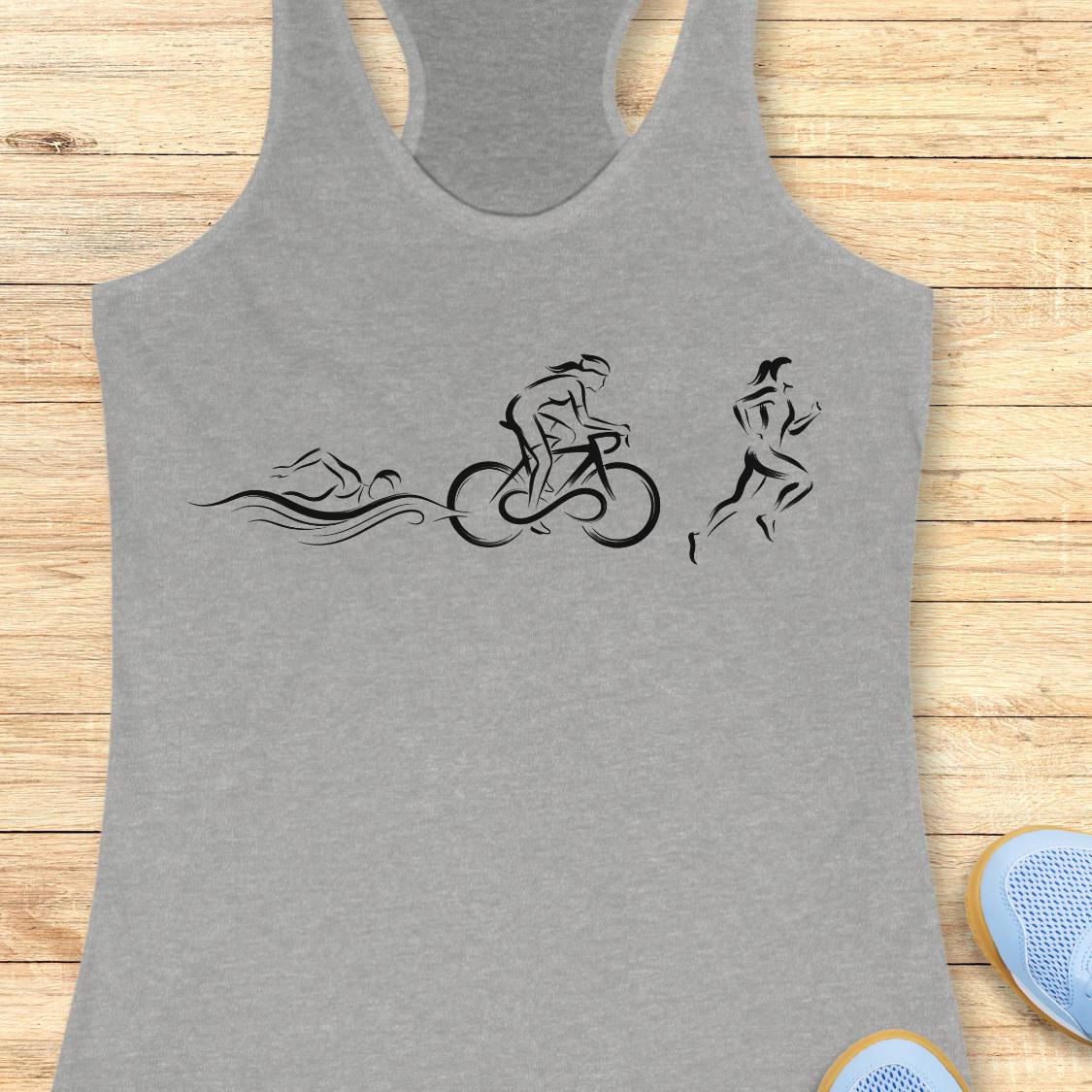 A heather gray racerback tank top with a black minimalist graphic of a swimmer, cyclist, and runner, laid flat on a light wood plank background with partial blue sneakers visible at the bottom right.