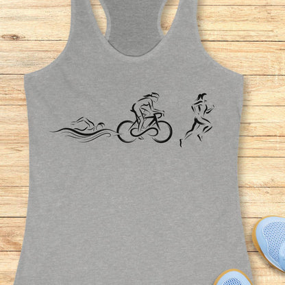A heather gray racerback tank top with a black minimalist graphic of a swimmer, cyclist, and runner, laid flat on a light wood plank background with partial blue sneakers visible at the bottom right.