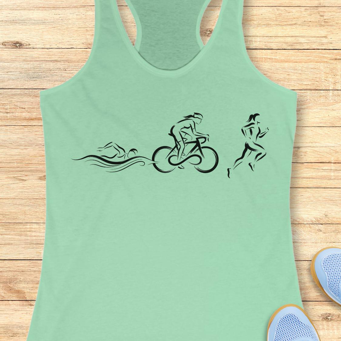 A mint green racerback tank top with a black line art graphic of a swimmer, cyclist, and runner, laid flat on a light wood plank background with partial blue sneakers visible at the bottom right.