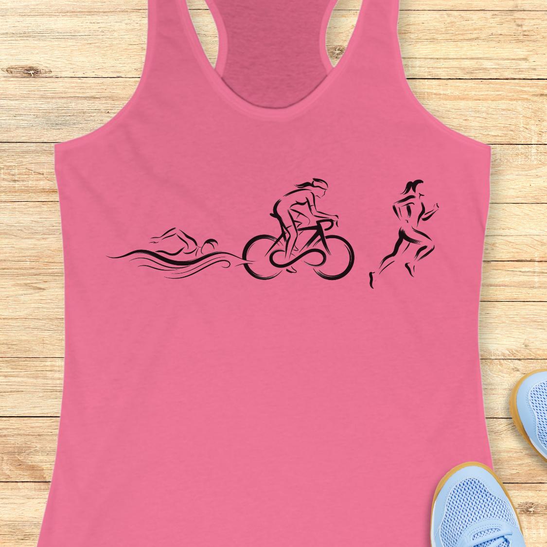 A pink racerback tank top laid flat on a light wood floor, featuring a black graphic of a swimmer, cyclist, and runner in sequence. Partial light blue sneakers are visible at the bottom right corners.