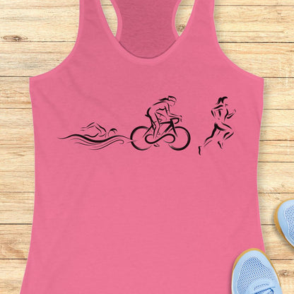 A pink racerback tank top laid flat on a light wood floor, featuring a black graphic of a swimmer, cyclist, and runner in sequence. Partial light blue sneakers are visible at the bottom right corners.