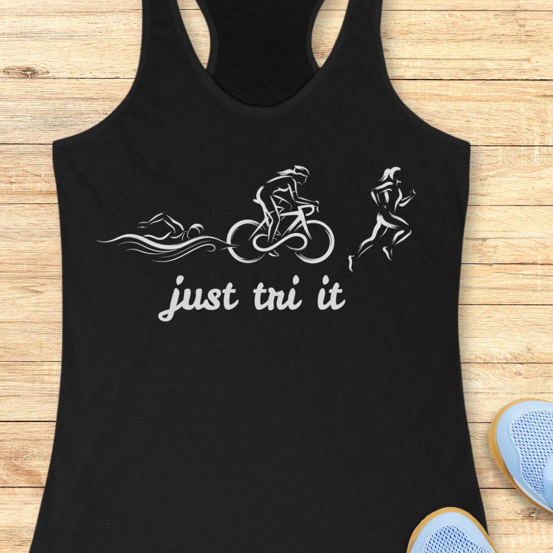A black racerback tank top laid flat on a light wood surface, featuring a white graphic of a swimmer, cyclist, and runner above the phrase 'just tn' it'. Partial blue sneakers are visible at the bottom right as styling props.