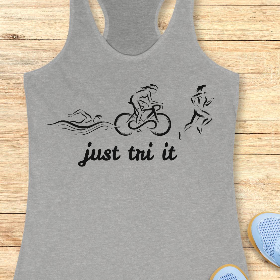 A heather gray racerback tank top with a black graphic of three athletes (swimmer, cyclist, runner) and the text 'just tri it' printed on the front, displayed flat on a light wood plank background.