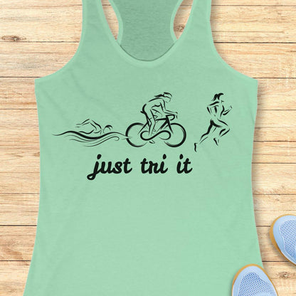A mint green racerback tank top with a black graphic of triathlon athletes and the text 'just tri it' on the front, displayed flat on a light wood plank background.
