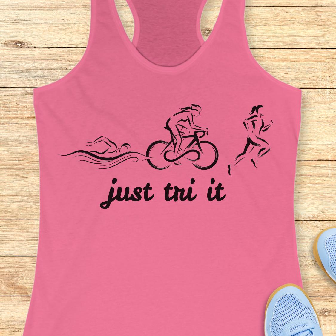 A pink racerback tank top laid flat on a light wood background, featuring a black graphic of a swimmer, cyclist, and runner above the words 'just tri it'. Partial blue sneakers are visible at the bottom right as styling props.