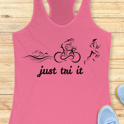 A pink racerback tank top laid flat on a light wood background, featuring a black graphic of a swimmer, cyclist, and runner above the words 'just tri it'. Partial blue sneakers are visible at the bottom right as styling props.