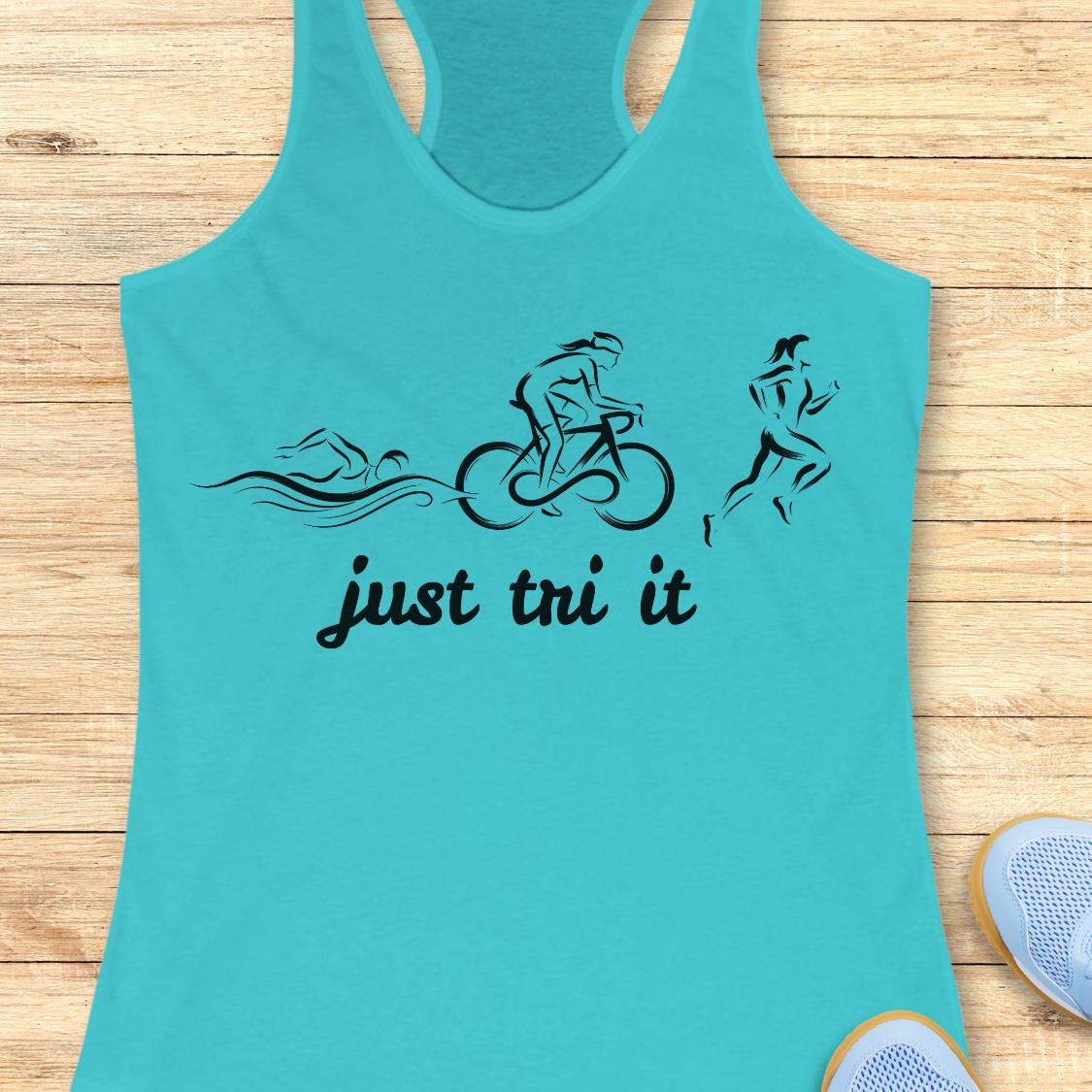 A turquoise racerback tank top laid flat on a light wood background, featuring a black graphic of a swimmer, cyclist, and runner above the words 'just tri it'. Partial light blue sneakers are visible at the bottom right corner.