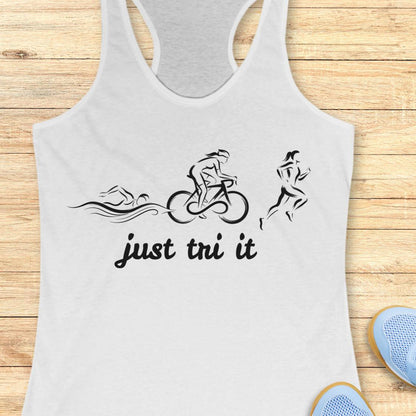 A white racerback tank top with a black graphic of a swimmer, cyclist, and runner above the text 'just tri it', displayed flat on a light wood background with blue sneakers partially visible in the corner.