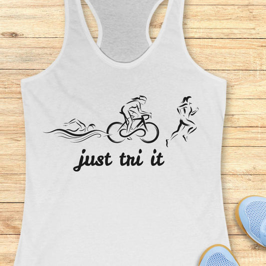 A white racerback tank top with a black graphic of a swimmer, cyclist, and runner above the text 'just tri it', displayed flat on a light wood background with blue sneakers partially visible in the corner.