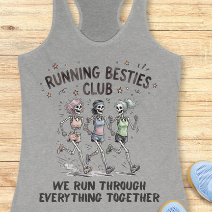 A heather gray racerback tank top laid flat on a light wood surface, featuring a printed graphic of three cartoon skeletons running together with the text 'RUNNING BESTIES CLUB' and 'WE RUN THROUGH EVERYTHING TOGETHER.' A partial blue shoe is visible at the lower right edge.