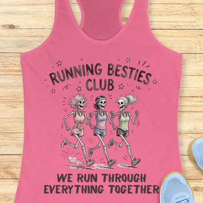 A pink racerback tank top laid flat on a light wood surface, featuring a graphic of three running skeletons with the text 'RUNNING BESTIES CLUB' and 'WE RUN THROUGH EVERYTHING TOGETHER.' Partial blue shoes are visible at the edges as styling props.