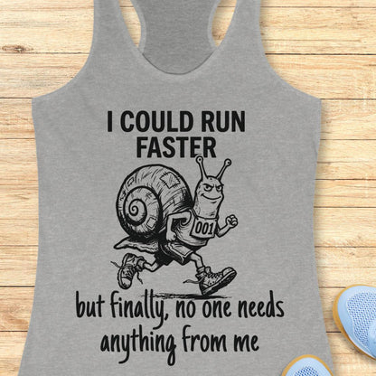 A heather gray racerback tank top laid flat on a light wood background, featuring a black graphic of a snail runner with the text 'I COULD RUN FASTER but finally, no one needs anything from me'.