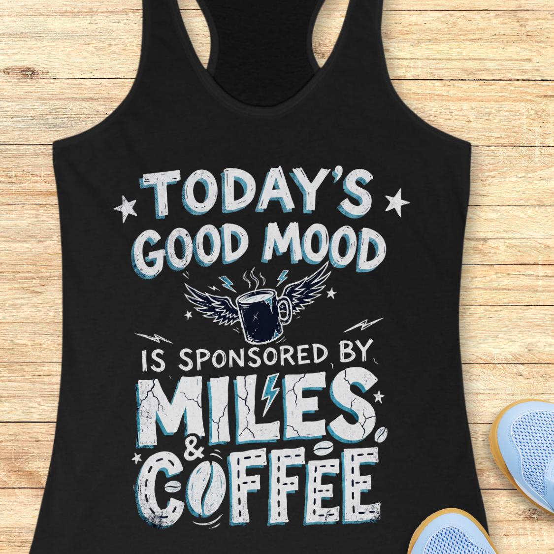 A black racerback tank top laid flat on a light wood background, featuring a distressed white and light blue graphic that reads 'TODAY'S GOOD MOOD IS SPONSORED BY MILES & COFFEE' with a winged coffee mug illustration. Partial blue shoes are visible at the bottom right as styling props.