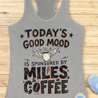 A heather gray racerback tank top laid flat on a light wood background, featuring a humorous graphic that reads 'TODAY'S GOOD MOOD IS SPONSORED BY MILES & COFFEE' with a coffee mug, wings, and lightning bolts. Partial blue shoes are visible at the bottom right as styling props.