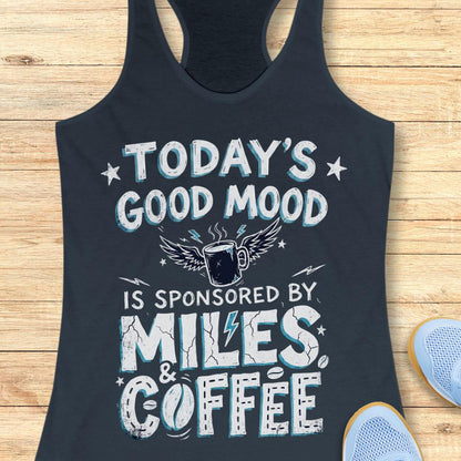 A racerback tank top laid flat on a light wood plank surface, featuring a humorous graphic that reads 'TODAY'S GOOD MOOD IS SPONSORED BY MILES & COFFEE' in distressed white and light blue lettering with a winged coffee mug illustration. Part of a blue shoe is visible at the bottom right.