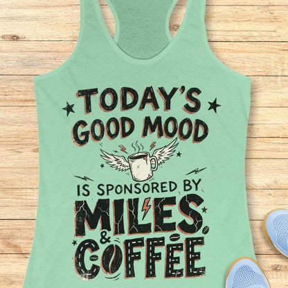 A mint green racerback tank top laid flat on a light wood background, featuring a graphic print that reads 'TODAY'S GOOD MOOD IS SPONSORED BY MILES & COFFEE' with a winged coffee mug illustration.