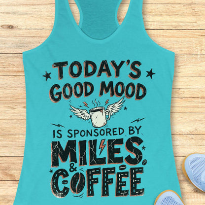 A turquoise racerback tank top laid flat on a light wood background, featuring a humorous graphic that reads 'TODAY'S GOOD MOOD IS SPONSORED BY MILES & COFFEE' with a winged coffee mug illustration. Part of a blue shoe is visible in the bottom right corner.