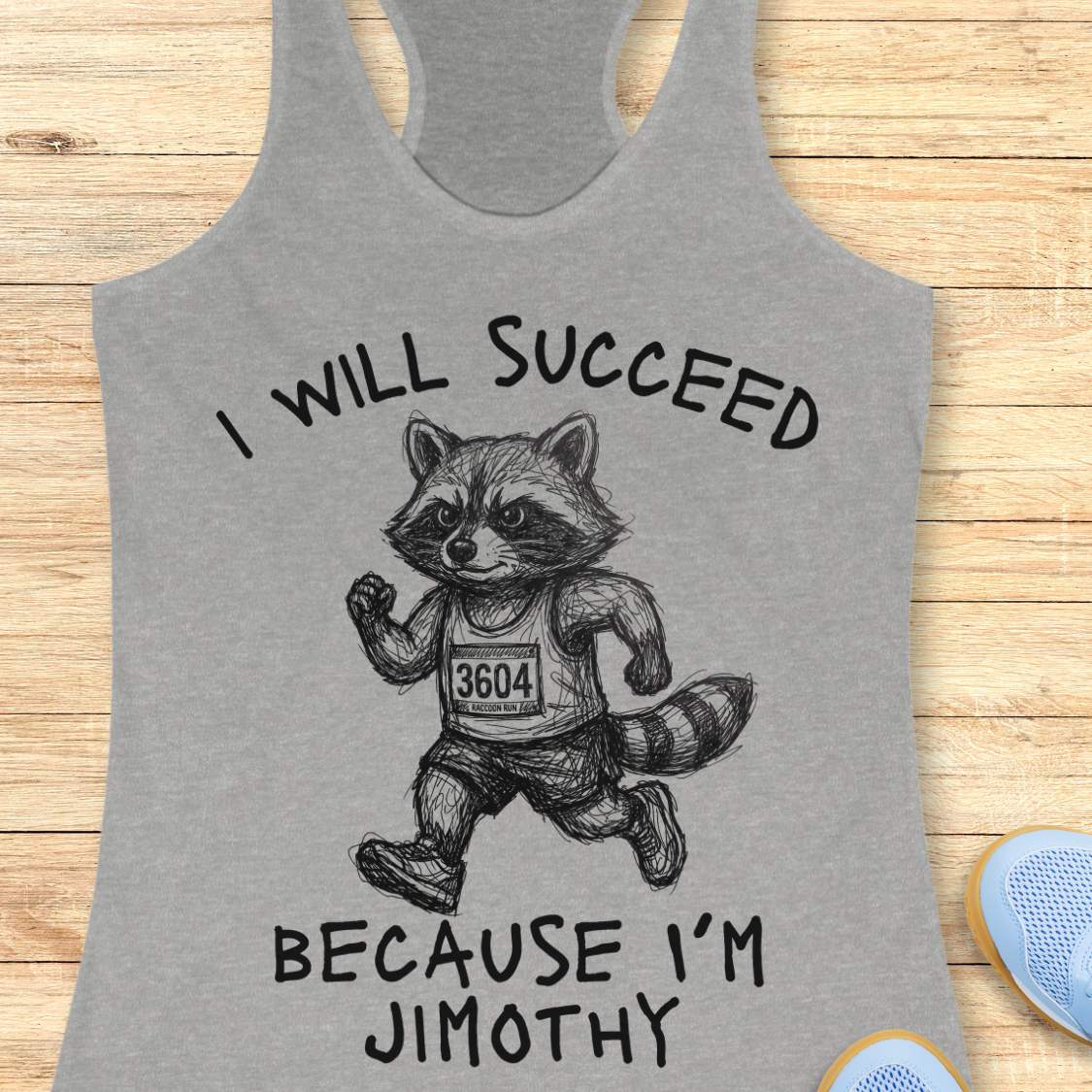 A heather gray racerback tank top laid flat on a light wood background, featuring a black line art graphic of a raccoon running with the text 'I WILL SUCCEED BECAUSE I'M JIMOTHY'.