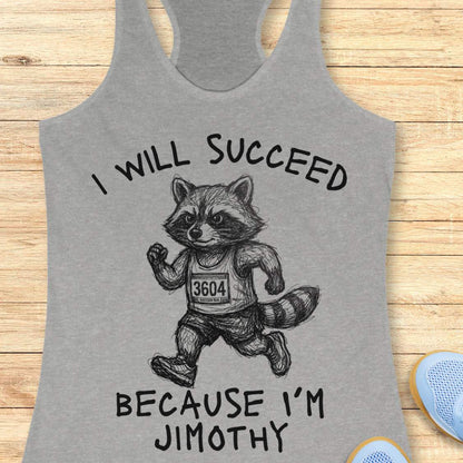 A heather gray racerback tank top laid flat on a light wood background, featuring a black line art graphic of a raccoon running with the text 'I WILL SUCCEED BECAUSE I'M JIMOTHY'.