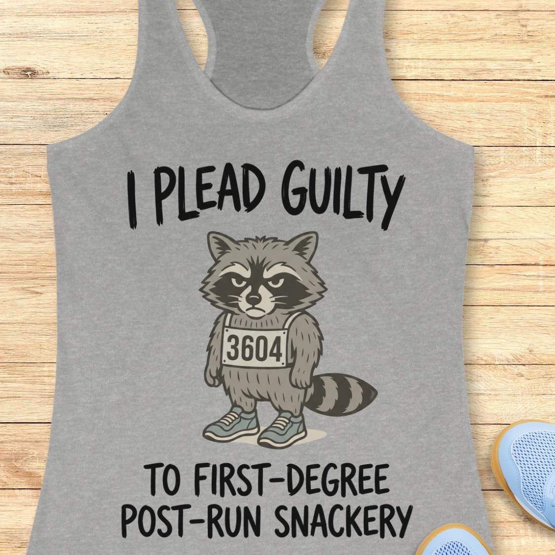 A heather gray racerback tank top laid flat on a light wood background, featuring a cartoon raccoon wearing a race bib numbered 3604 with the text 'I PLEAD GUILTY TO FIRST-DEGREE POST-RUN SNACKERY.' A partial blue shoe is visible at the bottom right.