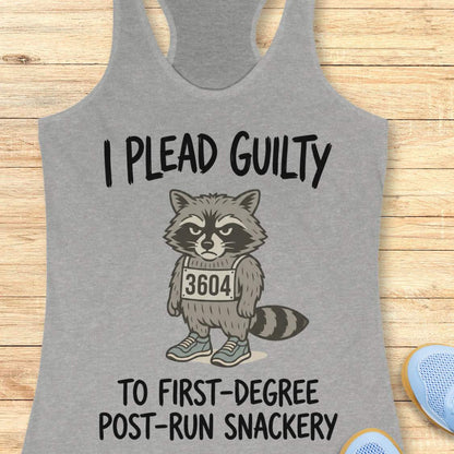A heather gray racerback tank top laid flat on a light wood background, featuring a cartoon raccoon wearing a race bib numbered 3604 with the text 'I PLEAD GUILTY TO FIRST-DEGREE POST-RUN SNACKERY.' A partial blue shoe is visible at the bottom right.