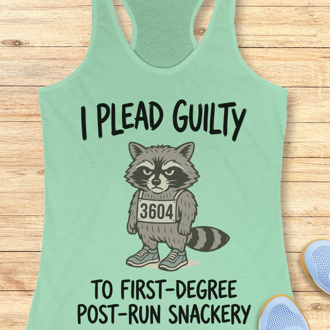 A mint green racerback tank top laid flat on a light wood background, featuring a cartoon raccoon wearing a mugshot-style number 3604 with the text 'I PLEAD GUILTY TO FIRST-DEGREE POST-RUN SNACKERY'. Partial blue shoes are visible at the bottom right.