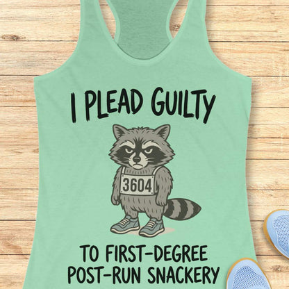 A mint green racerback tank top laid flat on a light wood background, featuring a cartoon raccoon wearing a mugshot-style number 3604 with the text 'I PLEAD GUILTY TO FIRST-DEGREE POST-RUN SNACKERY'. Partial blue shoes are visible at the bottom right.