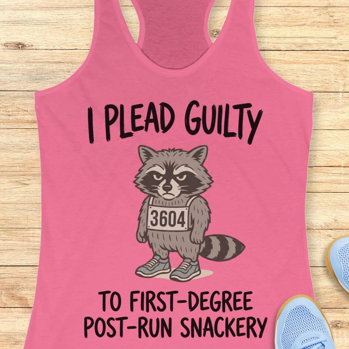 A pink racerback tank top laid flat on a light wood surface, featuring a cartoon raccoon wearing a race bib that reads 3604, with the text 'I PLEAD GUILTY TO FIRST-DEGREE POST-RUN SNACKERY.' Partial blue shoes are visible at the edges.