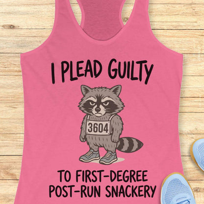 A pink racerback tank top laid flat on a light wood surface, featuring a cartoon raccoon wearing a race bib that reads 3604, with the text 'I PLEAD GUILTY TO FIRST-DEGREE POST-RUN SNACKERY.' Partial blue shoes are visible at the edges.