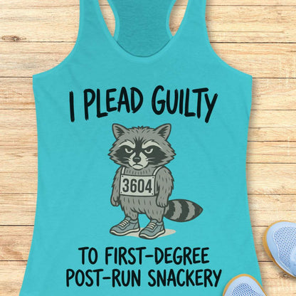 A turquoise racerback tank top laid flat on a light wood surface, featuring a printed graphic of a grumpy raccoon wearing a race bib numbered 3604, with the text 'I PLEAD GUILTY TO FIRST-DEGREE POST-RUN SNACKERY.' Partial blue shoes are visible at the bottom right as styling props.