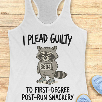 A white racerback tank top laid flat on a light wood background, featuring a cartoon raccoon wearing a race bib numbered 3604 with the text 'I PLEAD GUILTY TO FIRST-DEGREE POST-RUN SNACKERY.' Partial blue shoes are visible at the bottom right as styling props.