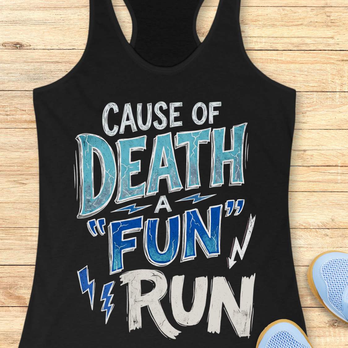 A black racerback tank top laid flat on a light wood background, featuring a distressed graphic that reads 'CAUSE OF DEATH A "FUN" RUN' in white and blue lettering. Part of a light blue shoe is visible at the bottom right as a styling prop.