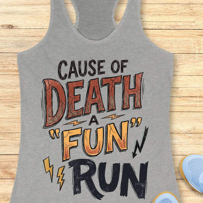 A heather gray racerback tank top with a graphic print reading 'CAUSE OF DEATH A "FUN" RUN' in black, orange, and brown lettering, laid flat on a light wood plank background with partial blue shoes visible at the edges.